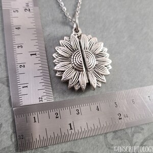 May include: Silver sunflower locket necklace with a chain. The locket is open and the two halves are separated. The locket is about 1 1/2 inches in diameter.
