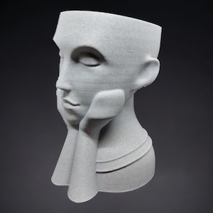 Peut inclure: Un pot en céramique blanche en forme de tête humaine avec les mains reposant sur le menton. Le pot a une surface texturée et un design simple.