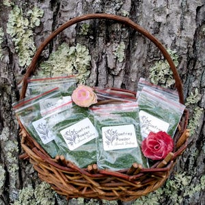 May include: A woven basket filled with small clear plastic bags containing green powder. Each bag has a white label with the text "Comfrey Powder" and a floral illustration. The basket is decorated with two artificial roses and sits against a textured tree trunk.