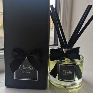 May include: A black box with a white label that says "Angel Bee Candles Diffuser". Next to the box is a clear glass diffuser bottle with a black bow and black reeds. The bottle has a white label that says "Angel Bee Candles Cinnamon and Orange".