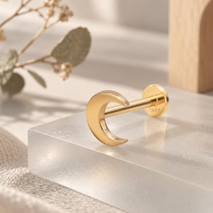 May include: A gold-coloured flat back stud earring featuring a crescent moon design. The post is clearly marked with the text 14K, indicating the metal purity. The jewellery piece has a polished, smooth finish and a circular base plate for secure wear.