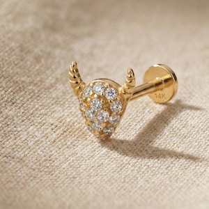 May include: A gold stud earring in the shape of a bull. The bull's head is embellished with multiple small, clear gemstones, and the horns are curved. The earring post is gold and marked with "14K".