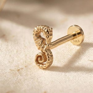 May include: A gold-coloured seahorse-shaped earring stud. The stud has a detailed seahorse design with a curved body and a flat, round backing. The earring is marked with "14KA".