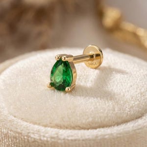 May include: A gold earring featuring a pear-shaped, bright green gemstone. The earring has a flat back and a post for piercing. The post is stamped with the text "14KA". The earring is set on a soft, cream-coloured surface.