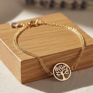 Puede incluir: Un brazalete dorado con un colgante de árbol de la vida, exhibido sobre un pequeño bloque de madera. El colgante es un diseño circular con una silueta detallada de árbol. El brazalete está hecho de delicados eslabones de cadena.