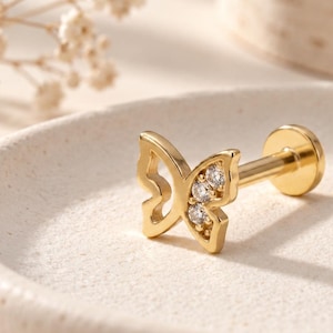 Könnte beinhalten: Ein goldfarbener Ohrstecker in Schmetterlingsform mit flachem Verschluss. Das Schmetterlingsdesign zeigt einen durchbrochenen Flügel auf der einen Seite und einen mit drei kleinen, klaren, rund geschliffenen Steinen besetzten Flügel auf der anderen.