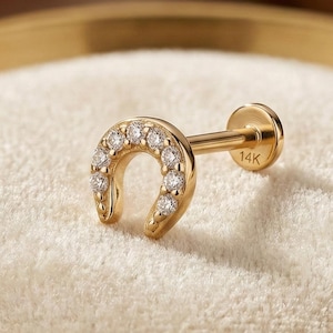 Peut inclure: Boucle d'oreille en forme de fer à cheval doré, ornée de petits pierres précieuses transparentes. L'image présente la boucle d'oreille sur une surface blanche texturée, dans un plat bordé d'or. La tige est marquée "14K".