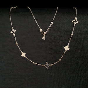 K&ouml;nnte beinhalten: Eine zarte goldene Halskette mit einer Reihe von vierbl&auml;ttrigen Blumenanh&auml;ngern und kleinen Perlenakzenten. Die Halskette hat eine verstellbare Kette und einen kleinen, tropfenf&ouml;rmigen Anh&auml;nger. Der Schmuck wird auf einer schwarzen Oberfl&auml;che pr&auml;sentiert.