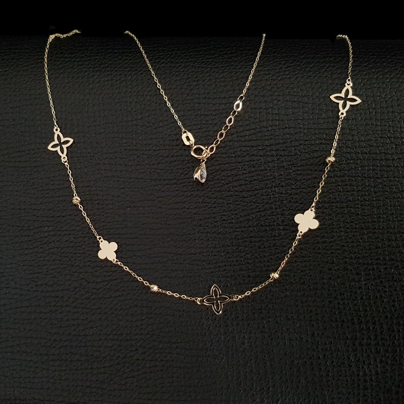 K&ouml;nnte beinhalten: Eine zarte goldene Halskette mit einer Reihe von vierbl&auml;ttrigen Blumenanh&auml;ngern und kleinen Perlenakzenten. Die Halskette hat eine verstellbare Kette und einen kleinen, tropfenf&ouml;rmigen Anh&auml;nger. Der Schmuck wird auf einer schwarzen Oberfl&auml;che pr&auml;sentiert.