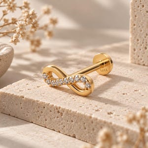 May include: A gold labret stud earring featuring an infinity symbol design adorned with small clear stones. The flat back post is clearly stamped with the text 14KA, indicating the metal composition. The jewelry piece has a polished finish.