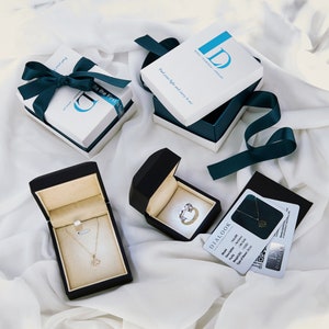 Puede incluir: Dos cajas de joyer&iacute;a de terciopelo negro con un collar y un anillo en su interior. Las cajas est&aacute;n sobre una superficie blanca con una caja de regalo blanca y azul y una caja de regalo blanca y turquesa. Las cajas de regalo tienen el texto "Be the First" y "Feel your light and carry it on." La imagen tambi&eacute;n incluye una tarjeta blanca con el texto "DIALOOK" y un c&oacute;digo QR.