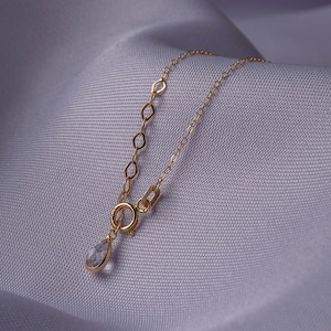K&ouml;nnte beinhalten: Eine zarte goldene Halskette mit einem tropfenf&ouml;rmigen Anh&auml;nger. Die Kette hat eine feine Kette mit kleinen, offenen Gliedern in der N&auml;he des Verschlusses. Der Anh&auml;nger ist ein klarer, facettierter Edelstein in einer goldenen Fassung, auf Lavendelstoff.