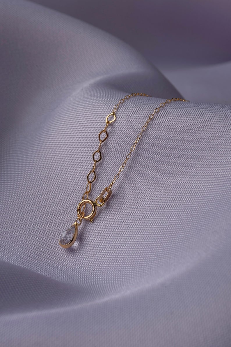 K&ouml;nnte beinhalten: Eine zarte goldene Halskette mit einem tropfenf&ouml;rmigen Anh&auml;nger. Die Kette hat eine feine Kette mit kleinen, offenen Gliedern in der N&auml;he des Verschlusses. Der Anh&auml;nger ist ein klarer, facettierter Edelstein in einer goldenen Fassung, auf Lavendelstoff.