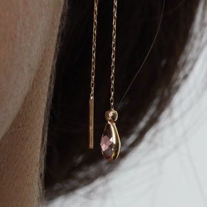May include: Gold-toned threader earrings. One earring features a slender bar, the other a chain with a teardrop-shaped pink gemstone pendant. The earrings are shown worn on an ear.