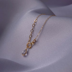Puede incluir: Un delicado collar de cadena dorada con un colgante de cristal transparente en forma de l&aacute;grima. El collar est&aacute; sobre un fondo de tela blanca.