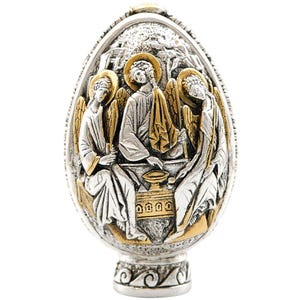 May include: An egg-shaped decorative object featuring a silver and gold-toned relief sculpture of three winged figures with halos seated around a table. The intricate metalwork displays detailed drapery and architectural elements on a pedestal base.