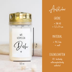 May include: A white candle with a gold lid and a sticker that reads "Wir vermissen Dich" (We miss you) in black lettering. The candle is 7.4 inches tall and 5.2 inches wide.
