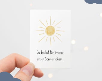 Può includere: Un cartoncino rettangolare bianco con un'illustrazione ad acquerello di un sole giallo con linee radianti. Sotto il sole, il testo nero recita: Du bleibst für immer unser Sonnenschein. Il cartoncino è tenuto contro uno sfondo chiaro.