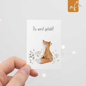 Könnte beinhalten: Eine rechteckige Karte mit einer Aquarellillustration eines Fuchses, der nach oben blickt. Der Fuchs ist orange und weiß, umgeben von weißen Gänseblümchen und grünen Blättern. Der Text "Du wirst geliebt!" steht oben.