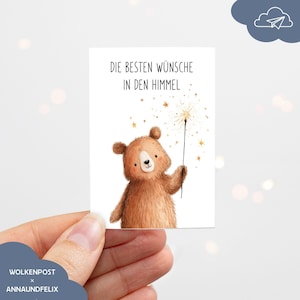 Könnte beinhalten: Eine rechteckige Grußkarte mit einer Illustration eines braunen Bären, der eine brennende Wunderkerze mit goldenen Sternen hält. Der Text oben lautet DIE BESTEN WÜNSCHE IN DEN HIMMEL in schwarzer Schrift auf weißem Grund.
