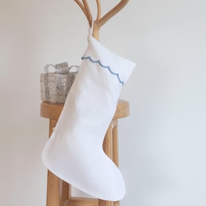 Puede incluir: Calcetín de Navidad de lino blanco con ribete azul festoneado colgado de un soporte de madera. Una caja de regalo plateada está en el soporte.