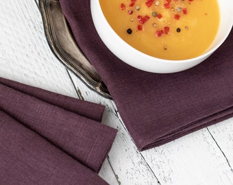May include: A white bowl of orange soup topped with red pepper flakes and black peppercorns. The bowl is sitting on a purple linen napkin on a white wooden table.