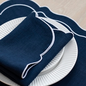 May include: A navy blue linen napkin with a white scalloped edge, folded and placed on a white plate with a scalloped edge. The napkin and plate are on top of a navy blue linen placemat with a white scalloped edge.