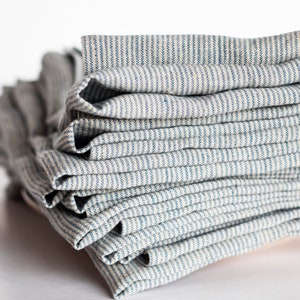May include: A stack of folded rectangular fabric napkins featuring a pattern of thin, vertical blue stripes on a light beige background. The fabric has a visible woven texture, and the napkins are neatly layered one on top of another.