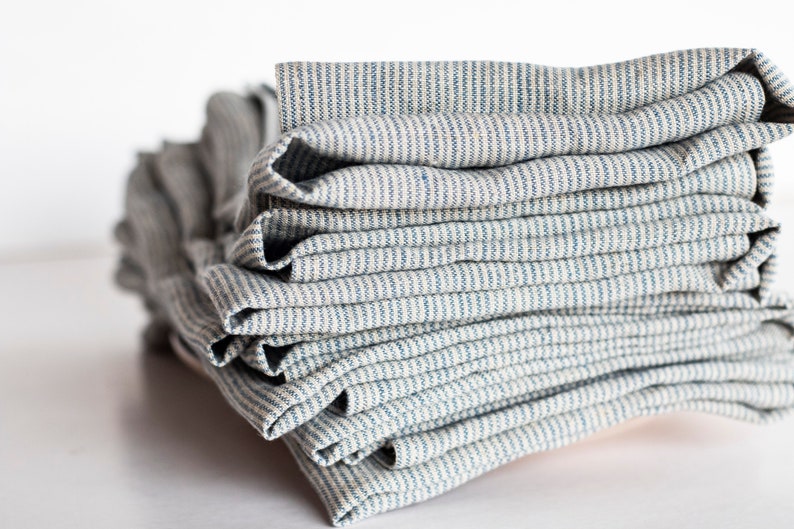 May include: A stack of folded rectangular fabric napkins featuring a pattern of thin, vertical blue stripes on a light beige background. The fabric has a visible woven texture, and the napkins are neatly layered one on top of another.