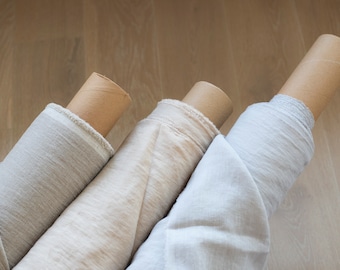May include: Three rolls of fabric in neutral colors: light gray, beige, and off-white. The fabric is rolled around cardboard tubes. The image is taken from a slightly elevated angle, showing the texture of the fabric.