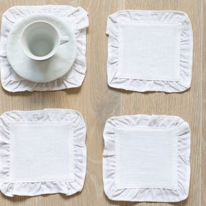 Op de afbeelding: Vier witte linnen onderzetters met gekrulde randen. Een onderzetter heeft een witte schotel en een wit theekopje erop.