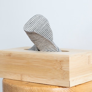Puede incluir: Una tapa de caja de pañuelos de madera con un pañuelo a rayas asomando por la abertura superior. La caja está hecha de bambú natural y tiene un color marrón claro.