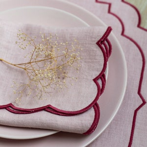 May include: A pink linen napkin with a scalloped edge and a burgundy trim. The napkin is folded and placed on a pink plate. A sprig of dried flowers is on top of the napkin.
