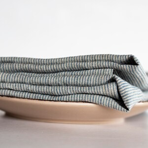 May include: A stack of folded blue and white striped linen napkins rests on a beige ceramic plate. The napkins have a textured appearance, and the stripes run horizontally across the fabric. The plate is round and has a simple, minimalist design.