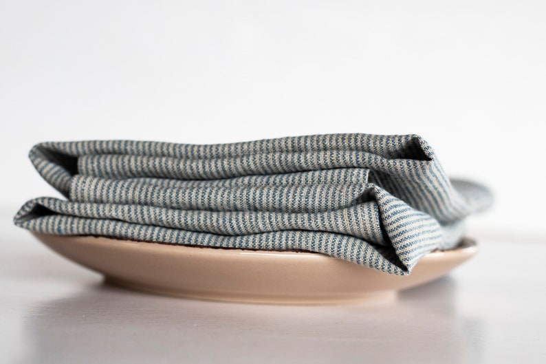 May include: A stack of folded blue and white striped linen napkins rests on a beige ceramic plate. The napkins have a textured appearance, and the stripes run horizontally across the fabric. The plate is round and has a simple, minimalist design.