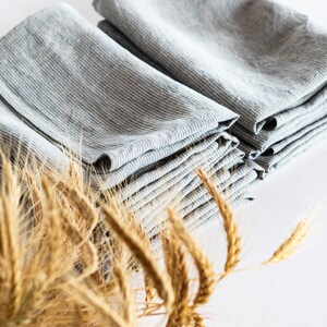 May include: Stacks of folded gray and white striped linen napkins. The napkins are arranged in neat piles, showcasing their texture and pattern. Dried wheat stalks are in the foreground, adding a rustic touch to the composition.