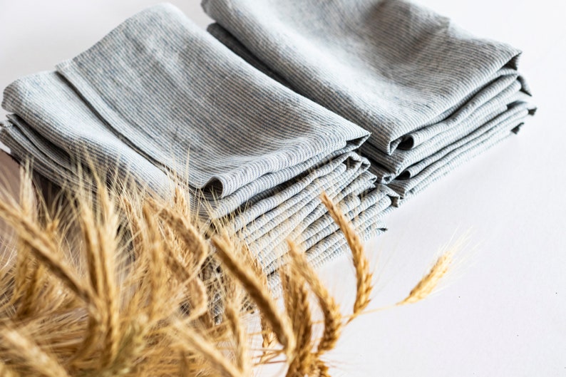 May include: Stacks of folded gray and white striped linen napkins. The napkins are arranged in neat piles, showcasing their texture and pattern. Dried wheat stalks are in the foreground, adding a rustic touch to the composition.