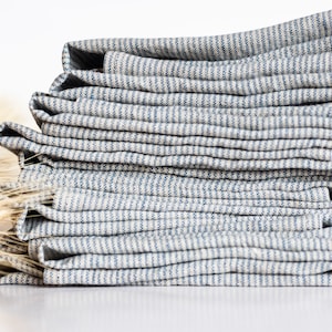 May include: A stack of folded, striped blue and white linen napkins. The napkins are neatly arranged, showcasing the fabric's texture and pattern. The image is set against a white background, with a few stalks of wheat in the background.