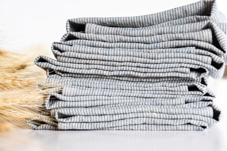 May include: A stack of folded, striped blue and white linen napkins. The napkins are neatly arranged, showcasing the fabric's texture and pattern. The image is set against a white background, with a few stalks of wheat in the background.