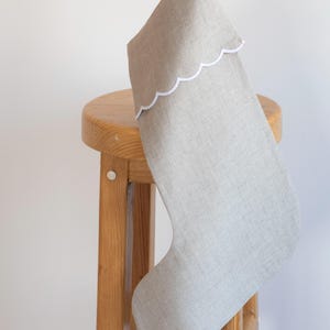 Könnte beinhalten: Ein grauer Weihnachtsstrumpf aus Leinen mit weißem, gewelltem Saum, der an einem Holzhocker hängt.