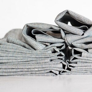 May include: Close-up of folded fabric with blue and white stripes. The fabric is stacked in a pile, with some sections rolled up. The material appears to be linen or a similar natural fiber, with a slightly textured surface.
