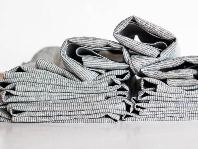 May include: Close-up of folded fabric with blue and white stripes. The fabric is stacked in a pile, with some sections rolled up. The material appears to be linen or a similar natural fiber, with a slightly textured surface.