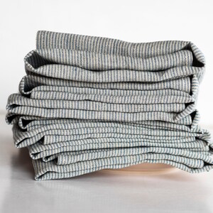 May include: A stack of folded, striped fabric napkins. The napkins have a light beige background with thin, vertical blue stripes. The fabric appears to be linen or a similar natural fiber. The napkins are neatly stacked on top of each other.
