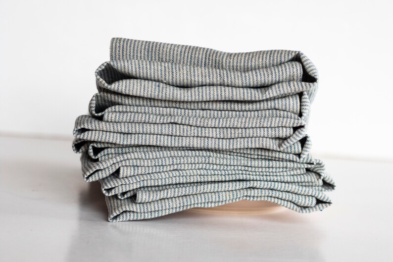 May include: A stack of folded, striped fabric napkins. The napkins have a light beige background with thin, vertical blue stripes. The fabric appears to be linen or a similar natural fiber. The napkins are neatly stacked on top of each other.