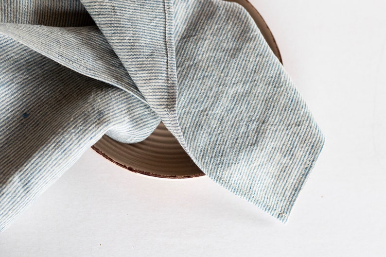 May include: A blue and white striped linen napkin is draped over a brown ceramic plate. The napkin's fabric has a textured appearance, with visible woven threads. The plate has a simple, rustic design.