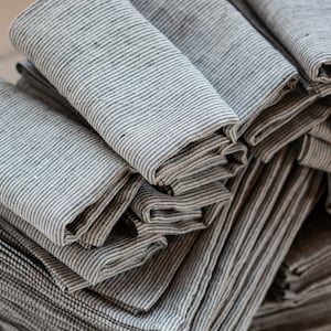 May include: A stack of folded gray and white striped linen napkins. The napkins are neatly arranged, showcasing the subtle texture and pattern of the fabric. The close-up shot highlights the natural, neutral tones and the quality of the material.