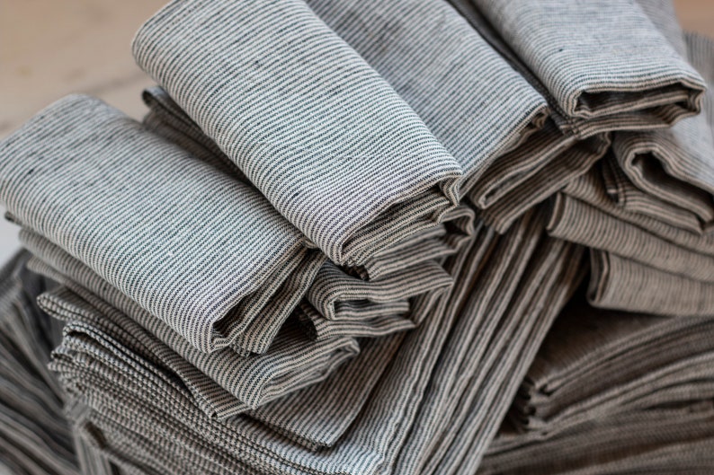 May include: A stack of folded gray and white striped linen napkins. The napkins are neatly arranged, showcasing the subtle texture and pattern of the fabric. The close-up shot highlights the natural, neutral tones and the quality of the material.