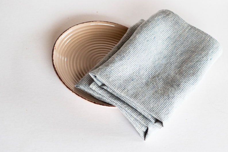 May include: A beige ceramic plate with a brown rim and a folded blue and white striped linen napkin. The plate has a ridged design. The napkin is folded and rests on the plate. The background is white.