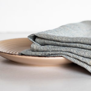 May include: A beige ceramic plate with a dark brown rim, topped with a folded blue and white striped linen napkin. The plate has a slightly textured surface. The items are set against a white background.