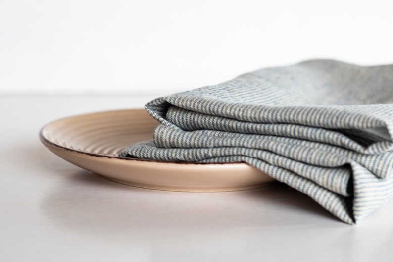 May include: A beige ceramic plate with a dark brown rim, topped with a folded blue and white striped linen napkin. The plate has a slightly textured surface. The items are set against a white background.